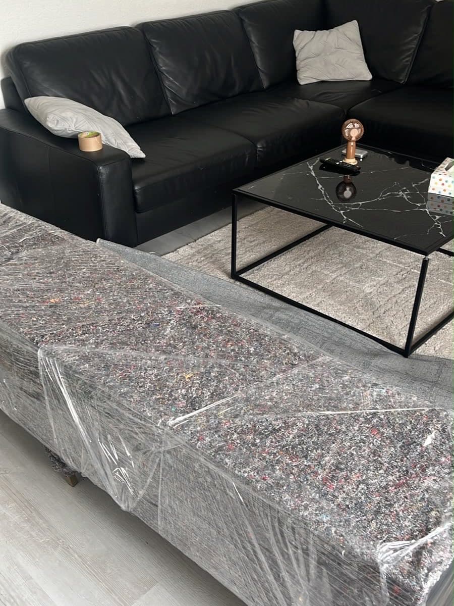 In einem Wohnzimmer ist ein Möbelstück mit Filzdecke und Folie abgedeckt, Sofa und Tisch stehen noch