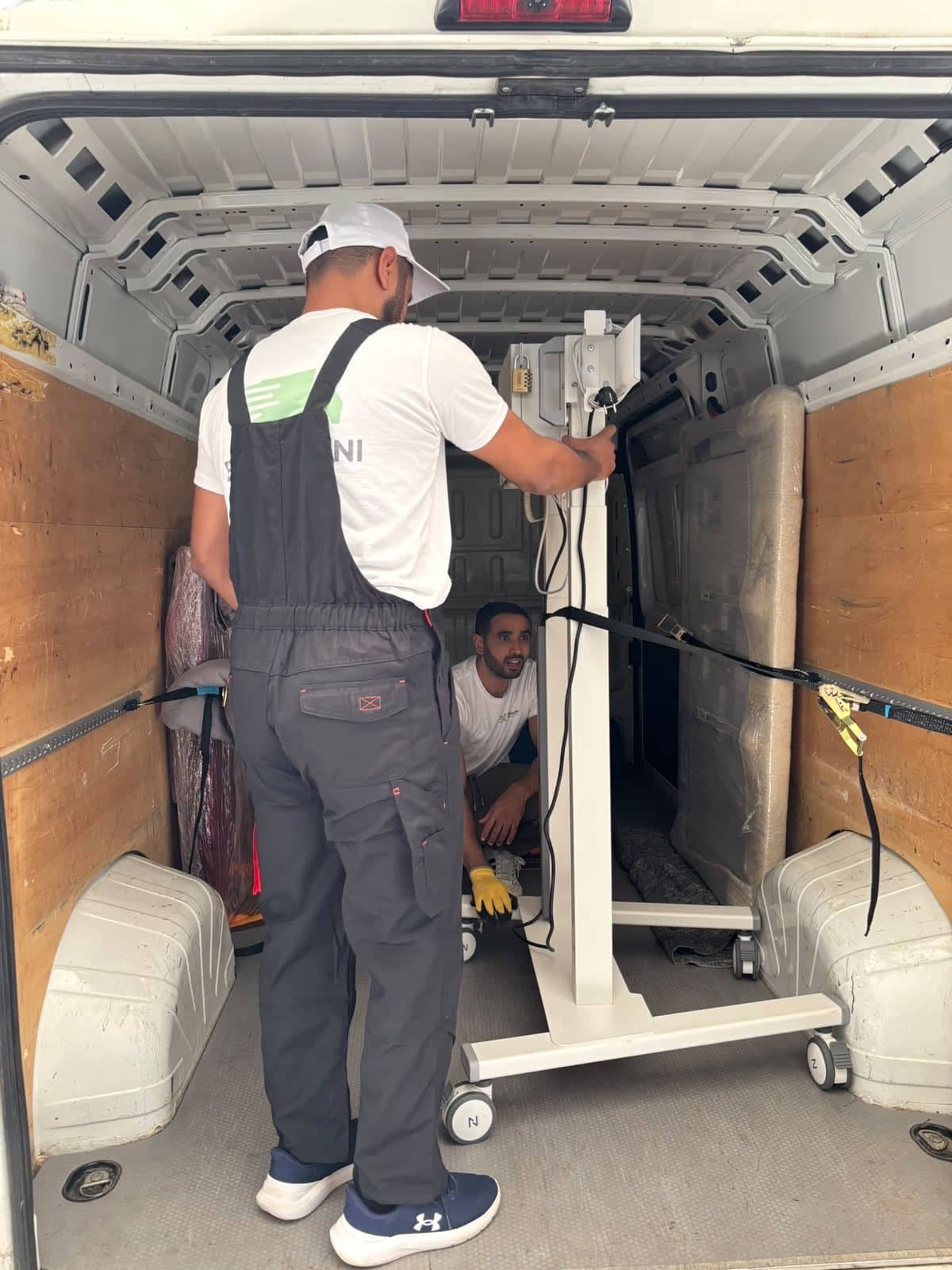 Zwei Mitarbeiter sichern ein Gerät auf Rollen mit Spanngurten im Laderaum eines Transporters