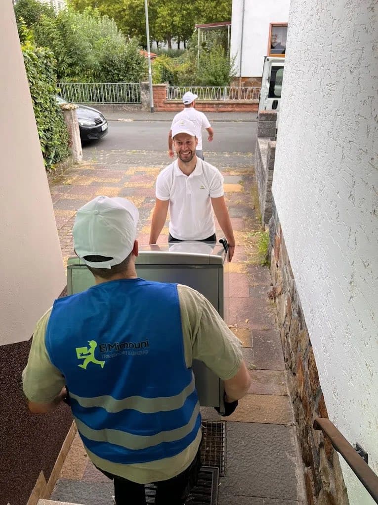 Zwei Mitarbeiter wuchten ein schweres Gerät auf einer Sackkarre über eine Außentreppe