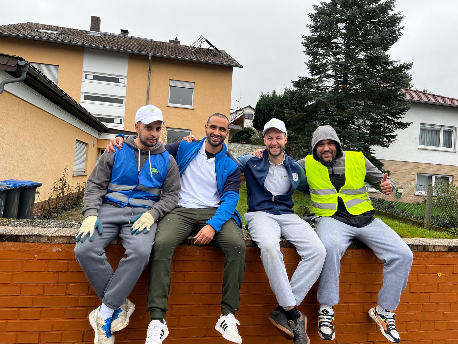 Four men sitting on a brick wall outside, smiling and posing with arms around each other, wearing casual sportswear and safety vests.