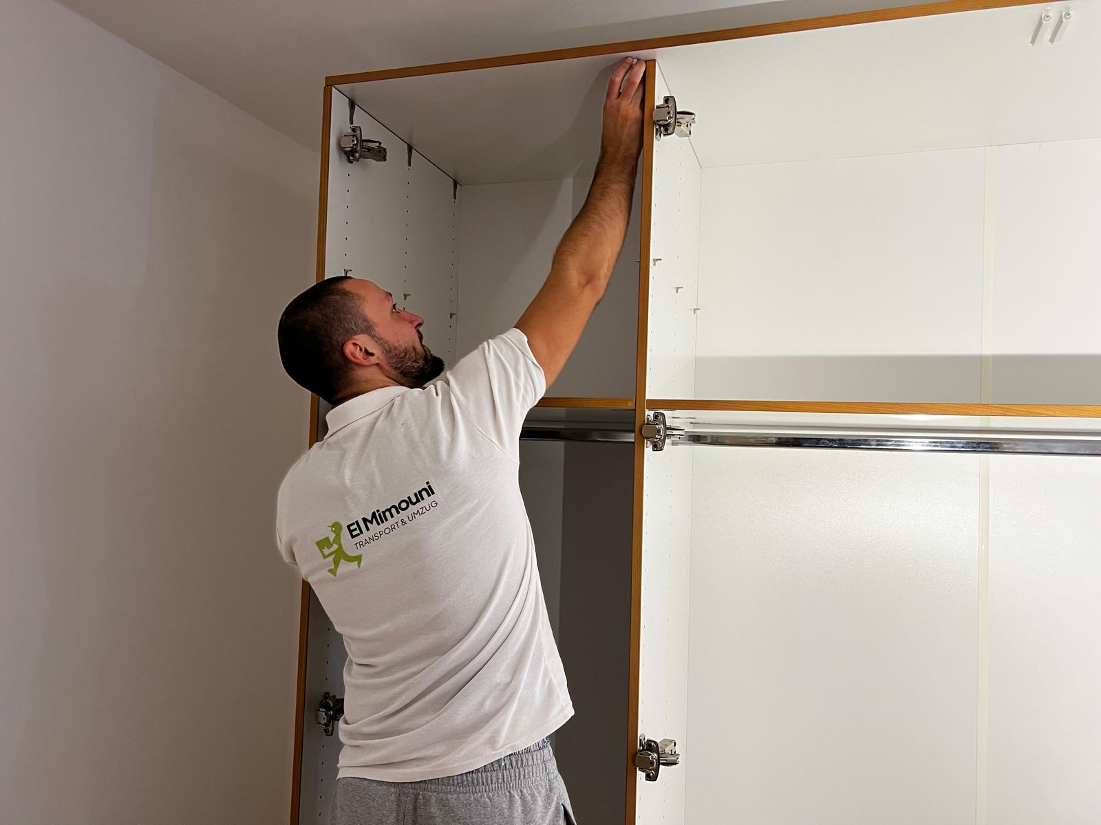 Man wearing a white shirt assembling or adjusting a white wardrobe cabinet inside a room.